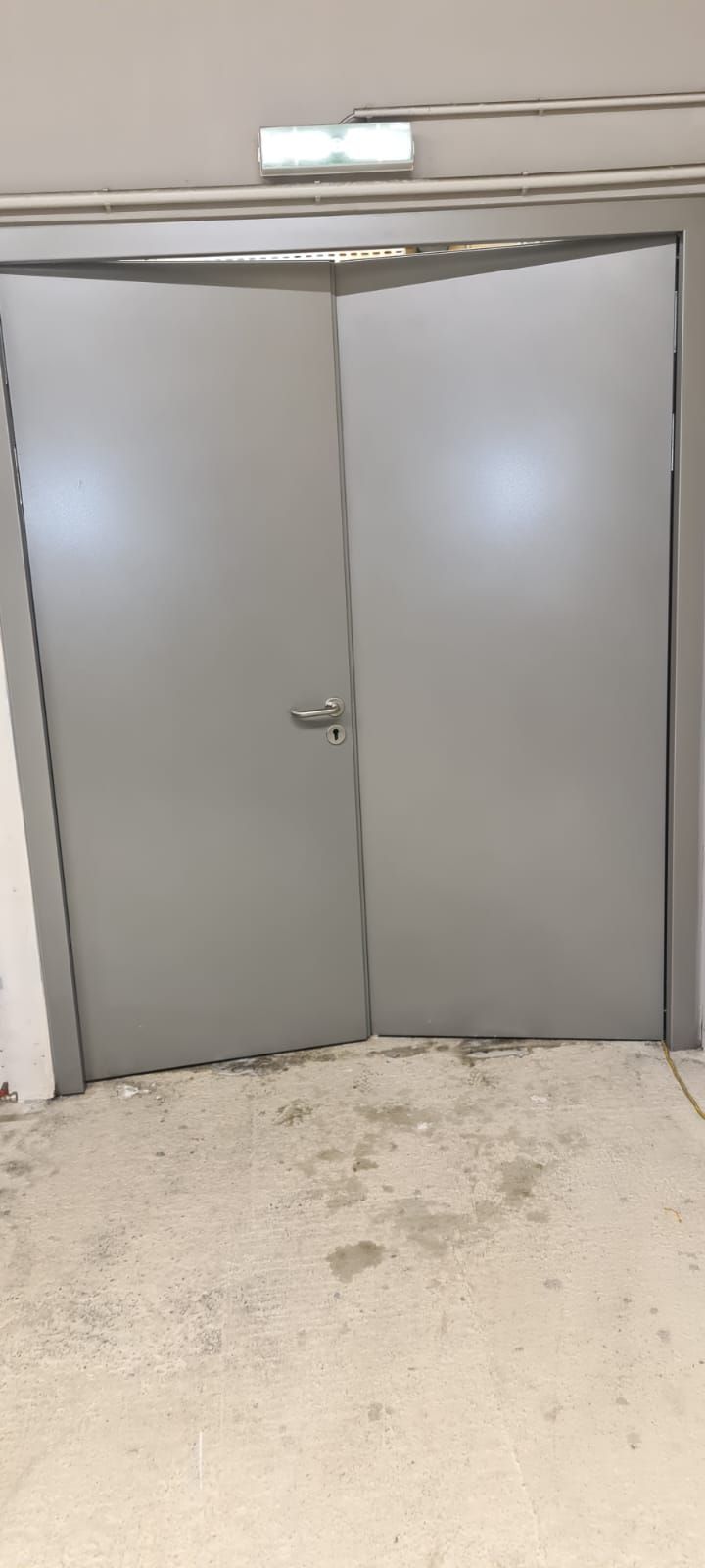 Graue Doppeltüren in einem Gebäude mit silbernem Rahmen, Griff und Notausgangsschild. Der Boden ist hell und fleckig.