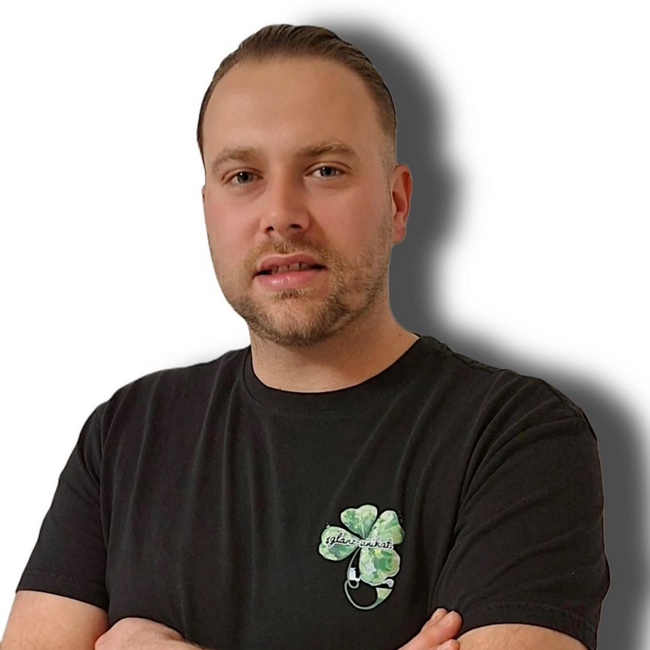 Mann in schwarzem T-Shirt mit vierblättrigem Kleeblattmuster, der nach vorne blickt und leicht lächelt.