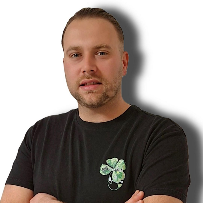 Mann in schwarzem T-Shirt mit vierblättrigem Kleeblattmuster, der nach vorne blickt und leicht lächelt.