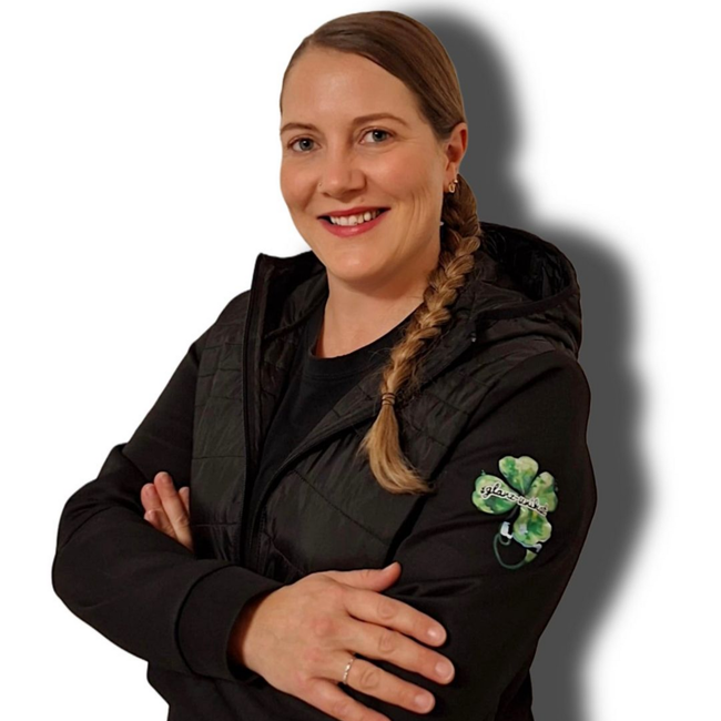 Frau in schwarzer Jacke mit geflochtenem Haar, die Arme verschränkt, lächelt. Kleeblatt-Aufnäher auf dem Ärmel.