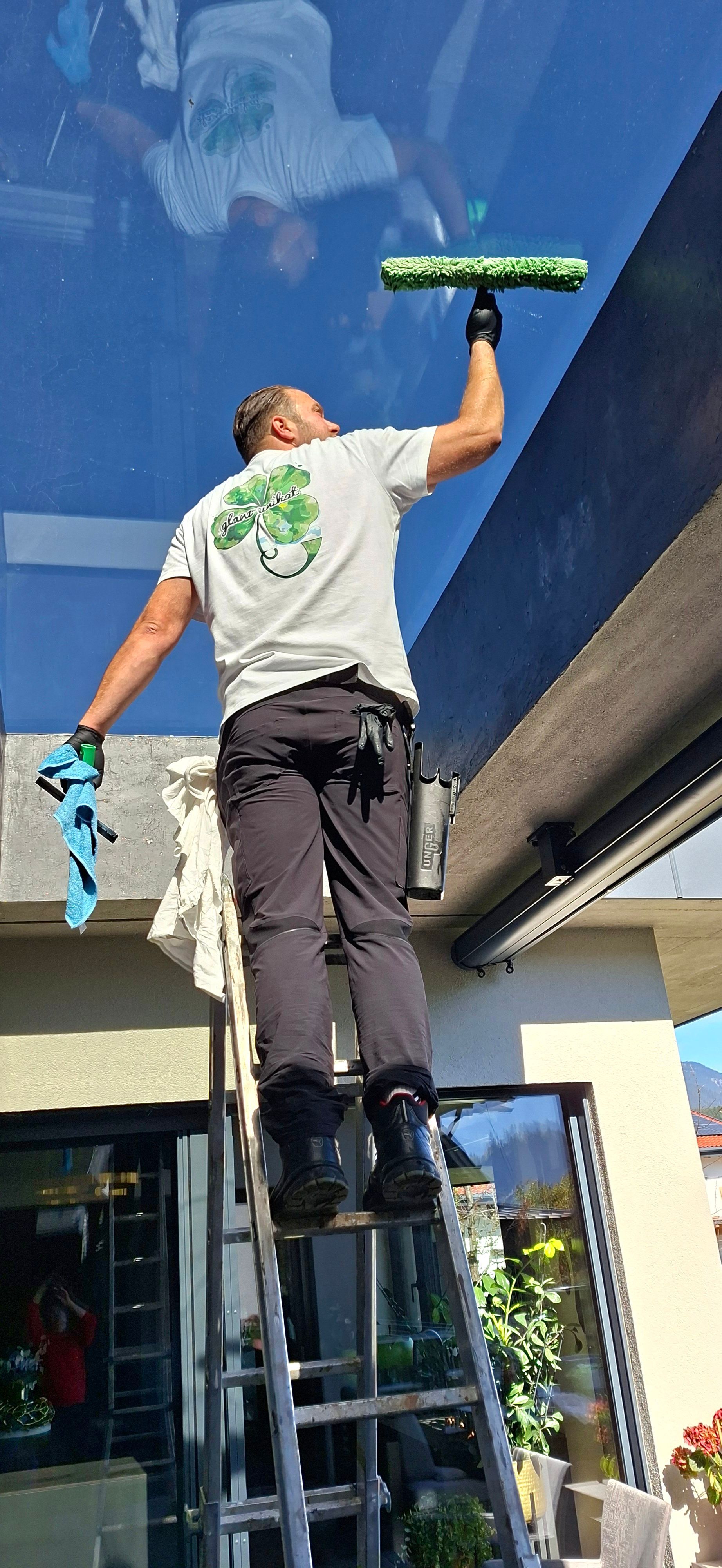 Ein Fensterputzer auf einer Leiter reinigt ein großes Glasfenster; er trägt ein weißes Hemd mit grünem Kleeblatt-Logo.