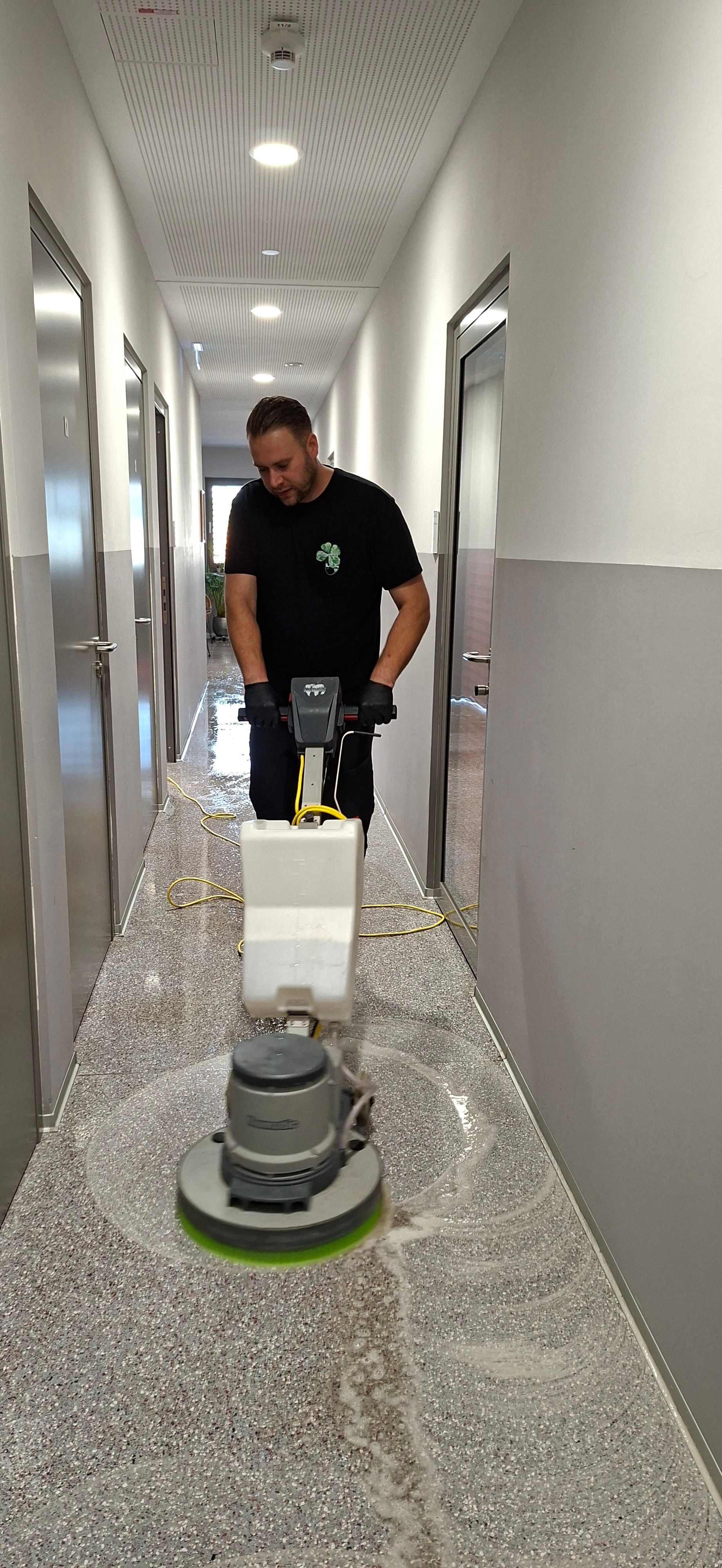 Ein Mann benutzt eine Bodenreinigungsmaschine in einem Flur mit grauen Wänden und Türen.