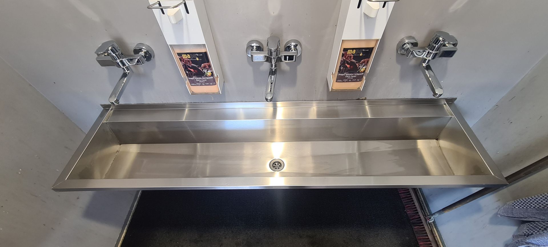 Industrielles Edelstahlspülbecken mit drei Wasserhähnen, an der Wand montiert.