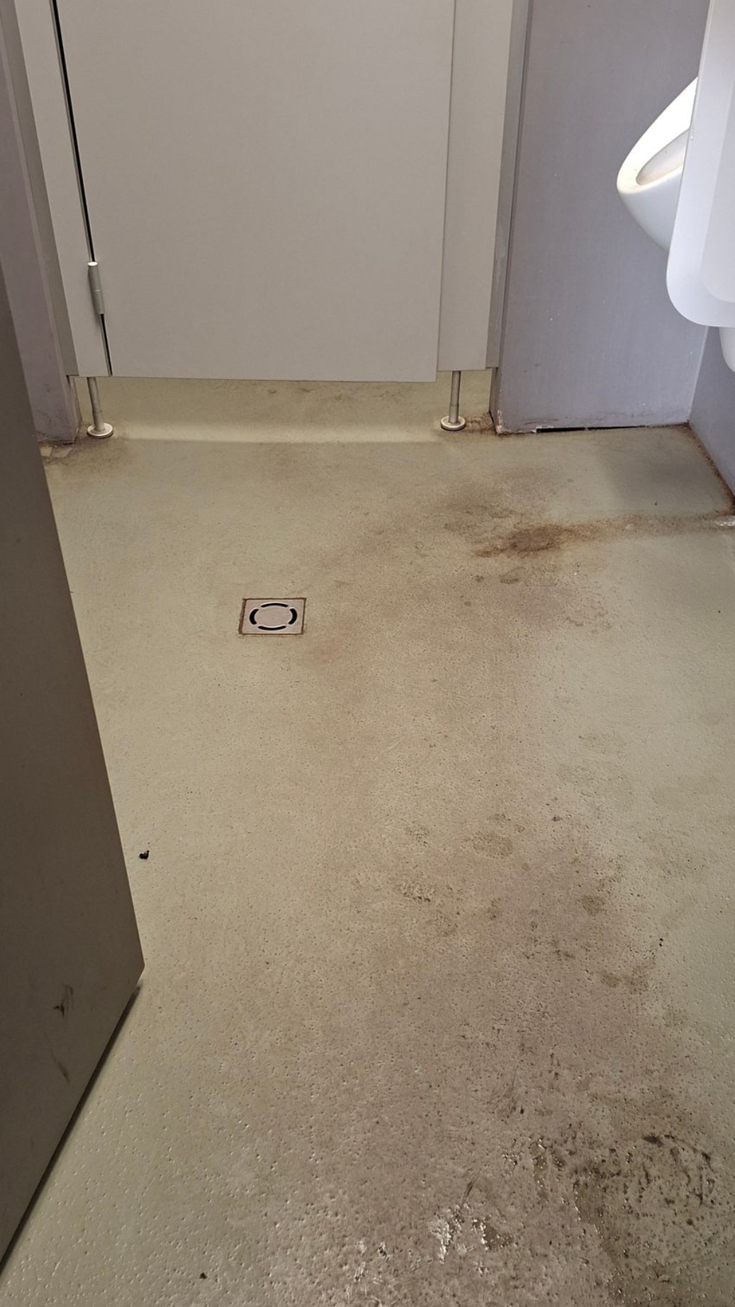Schmutziger Toilettenboden mit Flecken und einem Abfluss, neben einer Kabinentür und einem Urinal.