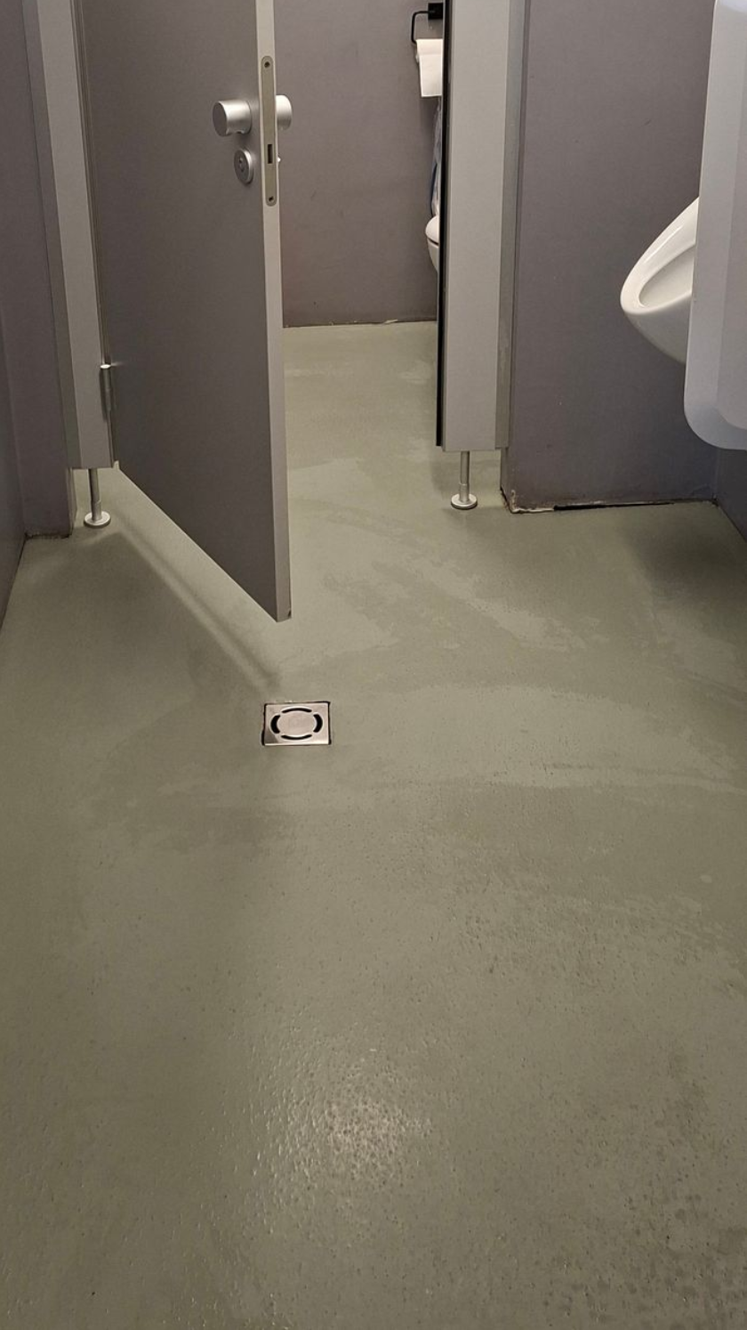 Innenansicht einer Toilettenkabine. Offene graue Tür, graue Wände, weißes Urinal sichtbar. Grauer Boden.