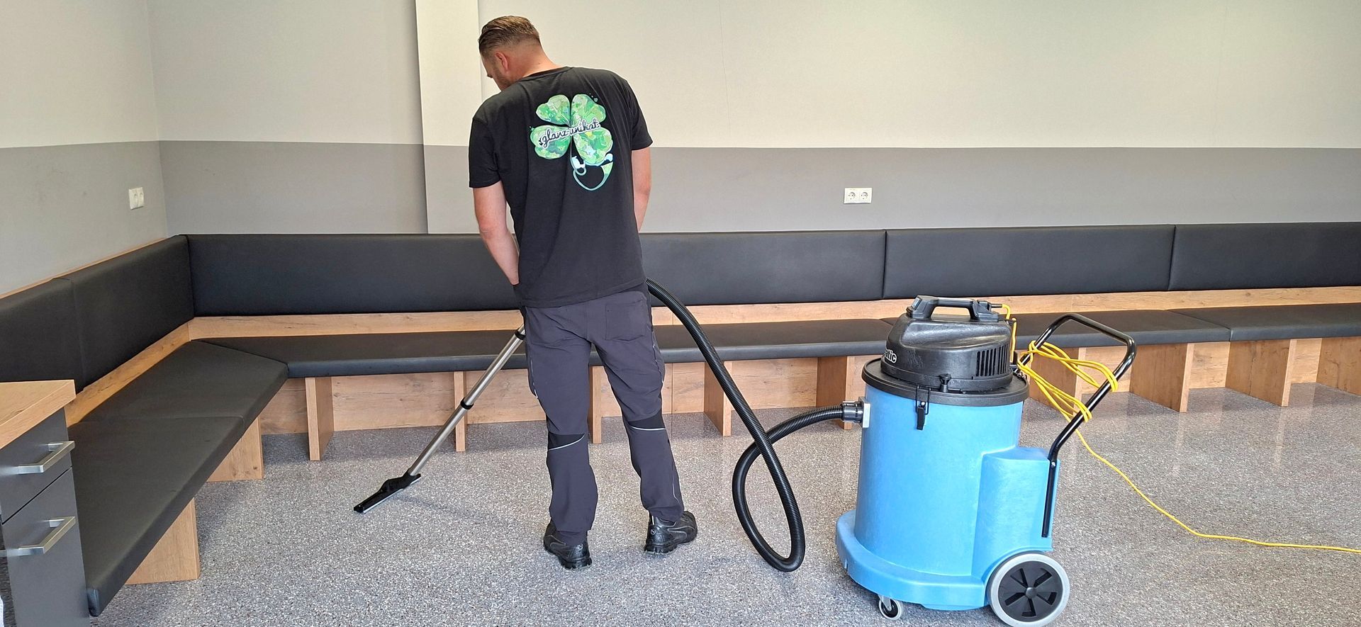 Eine Person saugt den Boden in der Nähe einer langen, gepolsterten Bank. Der Staubsauger ist blau und hat Rollen.
