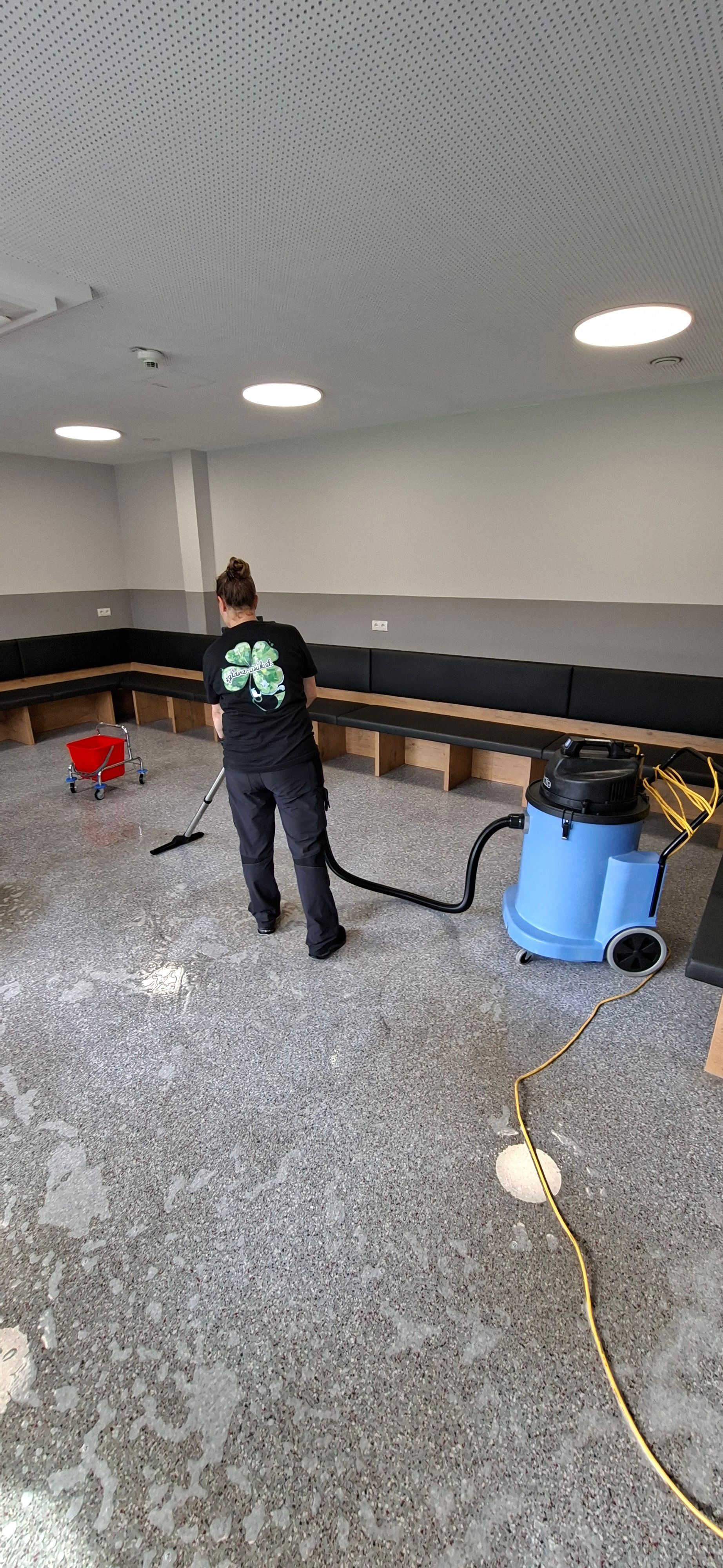 Eine Person saugt den Boden in einem Zimmer mit eingebauter Sitzbank. Blauer Staubsauger, helle Wände und Boden.