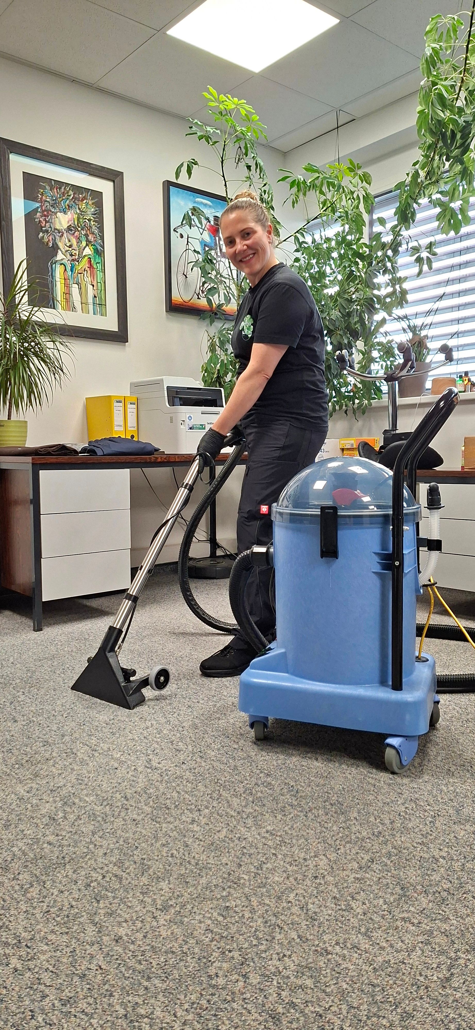 Eine Person in Schwarz reinigt mit einem blauen Staubsauger den Büroteppich.