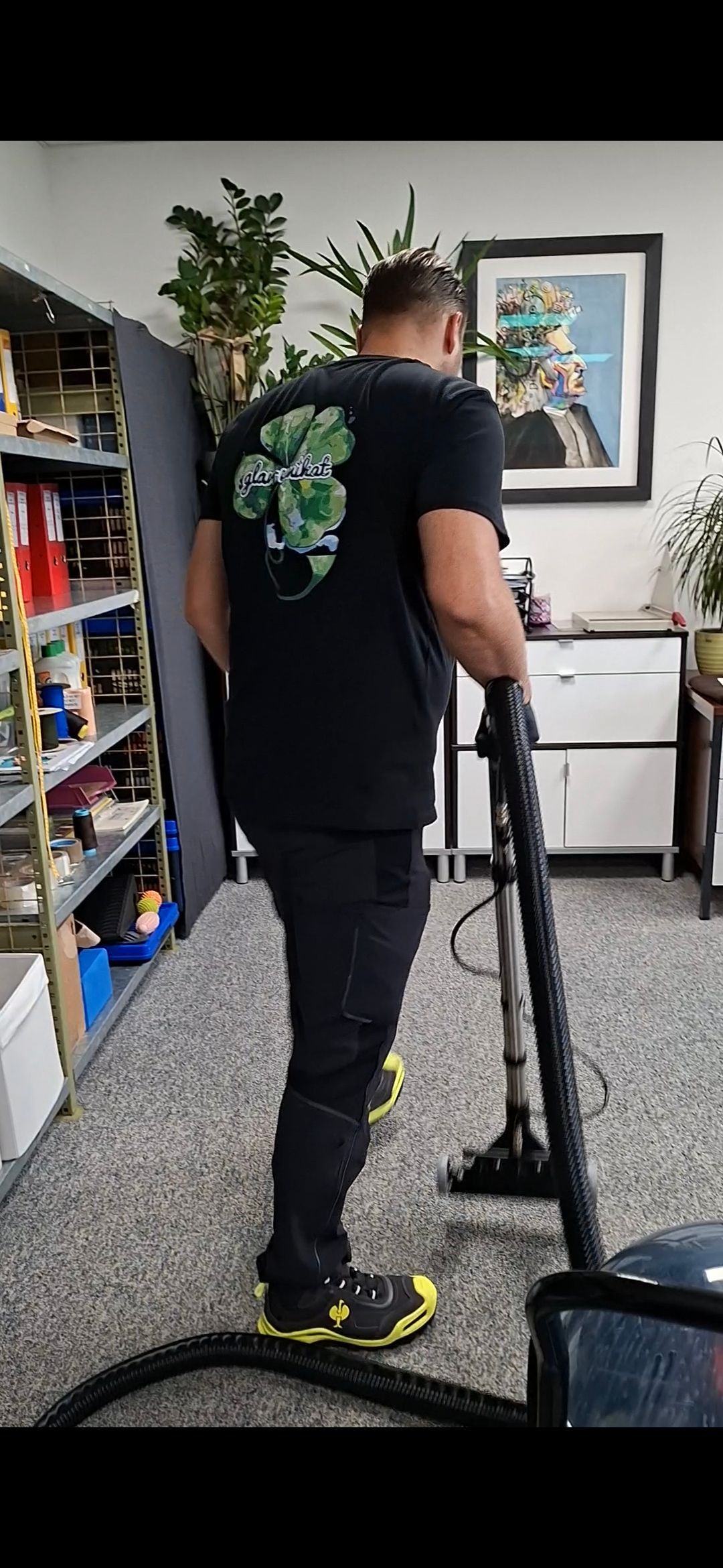 Eine Person saugt Staub in einem Zimmer mit Teppichboden. Die Person trägt ein schwarzes Hemd mit grünem Muster