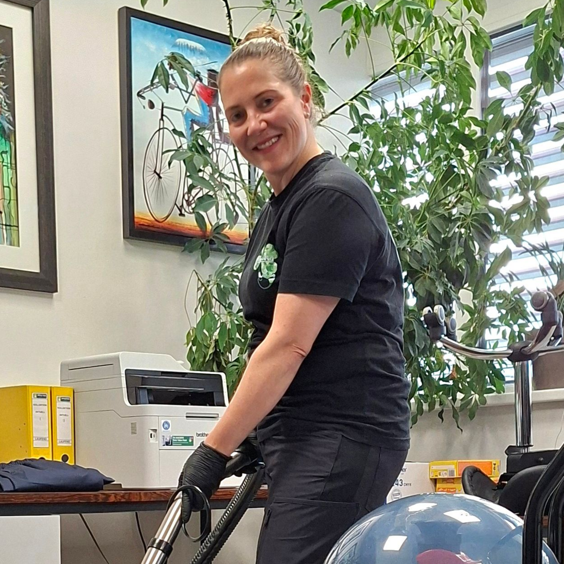 Frau saugt Staub in einem Büro, lächelt, trägt ein schwarzes Hemd und Handschuhe.