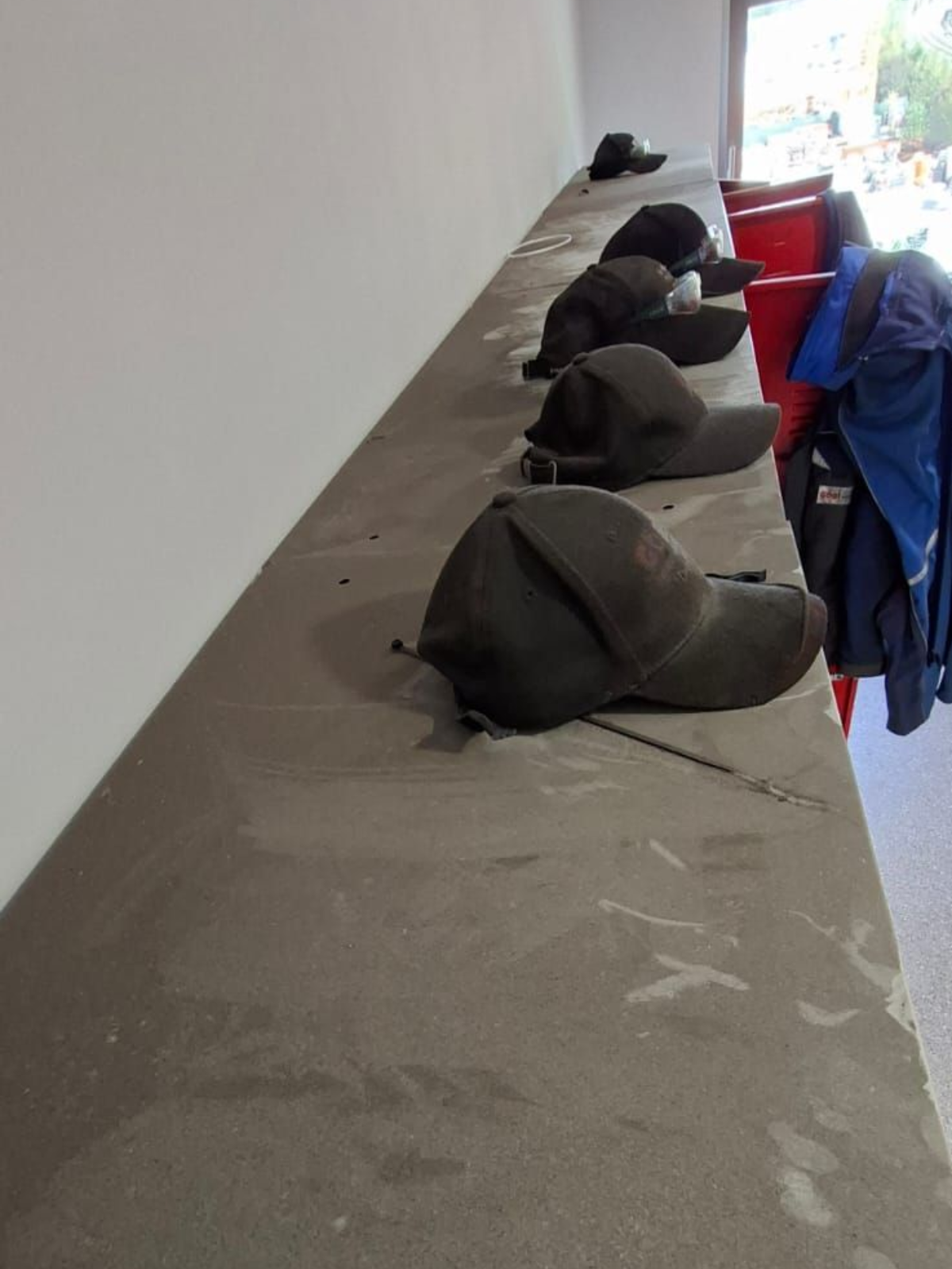 Auf einem staubigen, langen, grauen Regal neben einer weißen Wand stehen Baseballkappen.