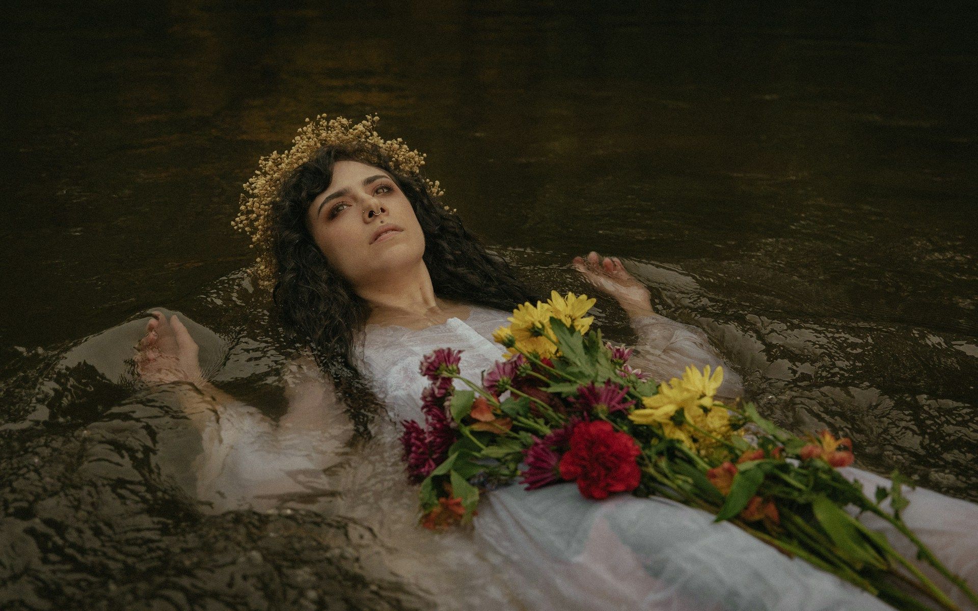 Woman floating in water, wearing a crown and white dress, with flowers; eyes closed, serene expression.