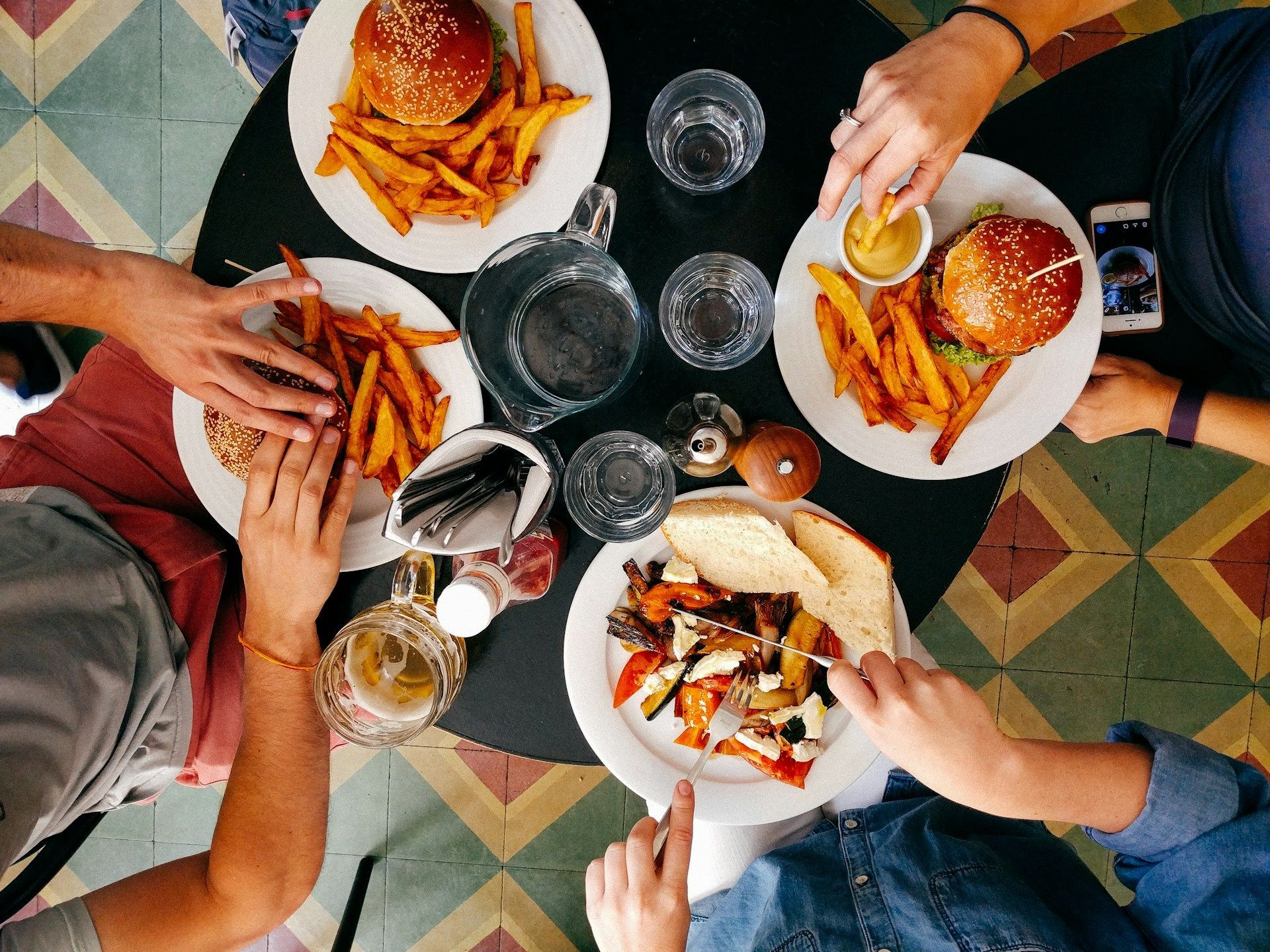 People eating burgers, fries, and other food at a table. Several hands reaching for food.