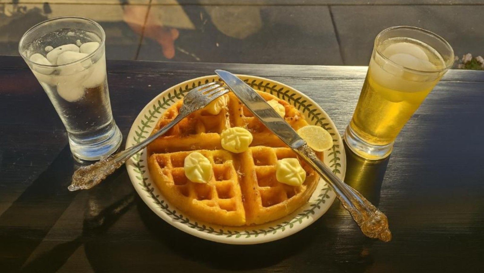 Waffle with butter, fork, and knife, with two drinks (one with ice) on a table.