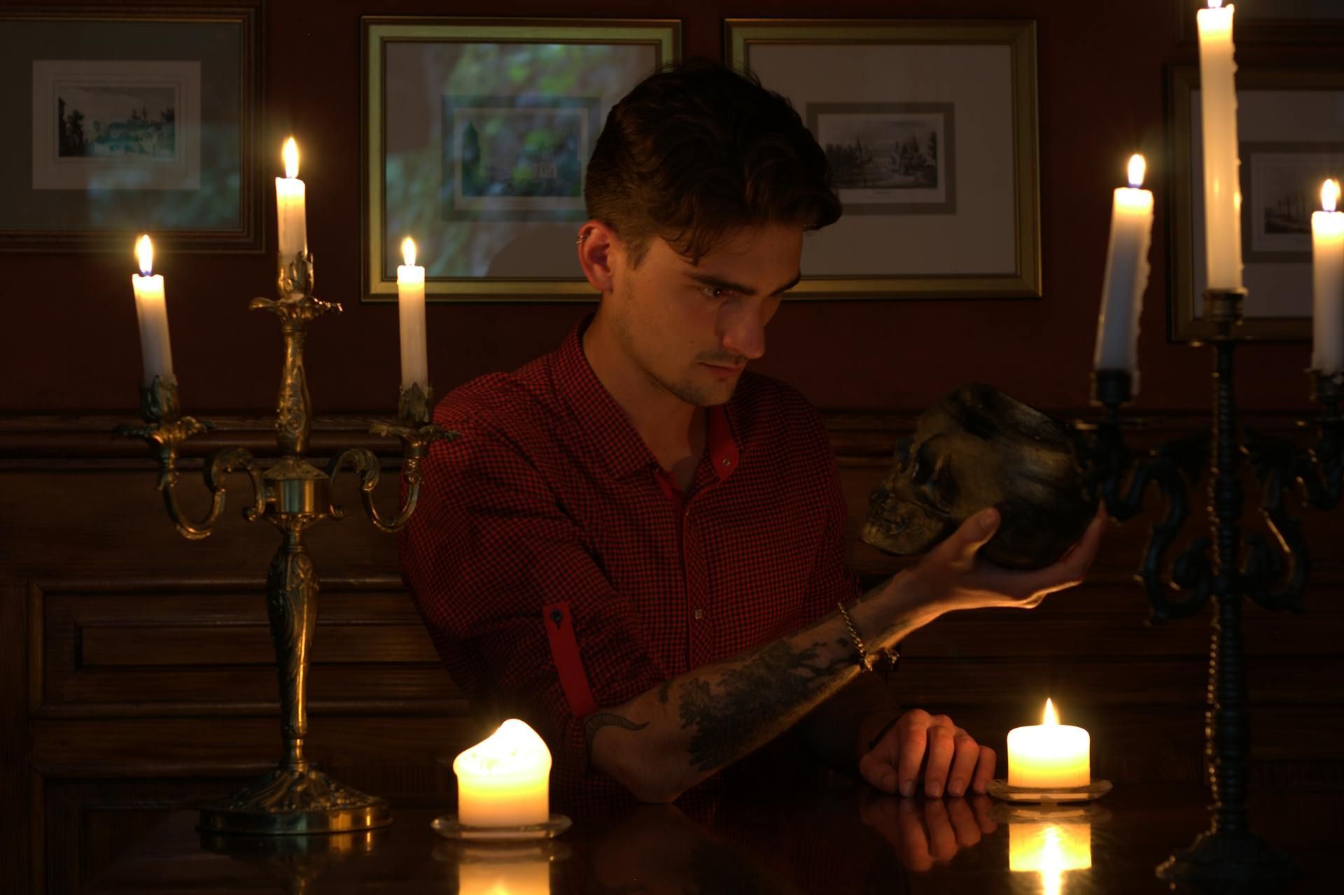 Man in red shirt holding skull, lit by candles in a room with framed art.