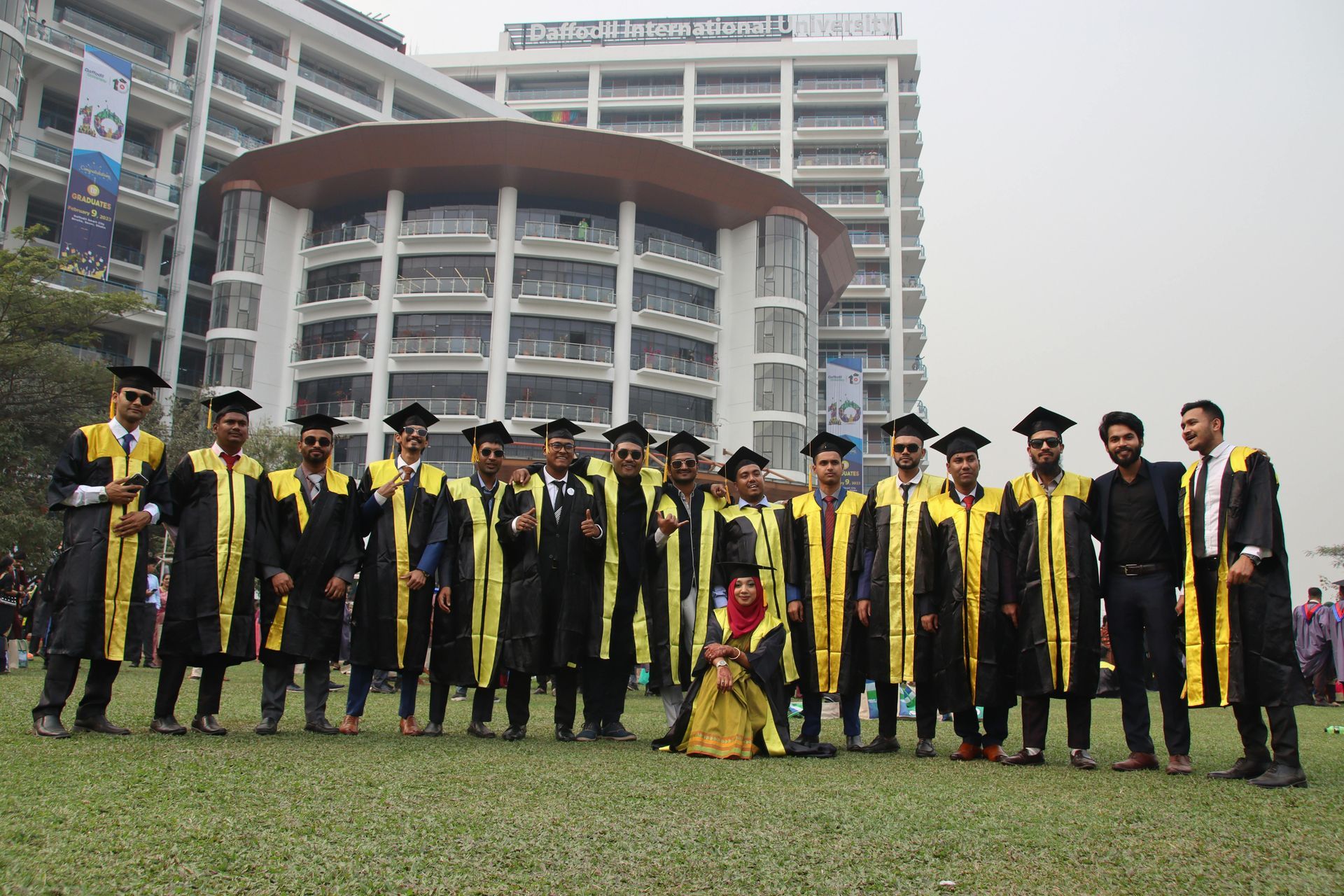 Group of graduates in black gowns with gold trim pose on a lawn, building in background.