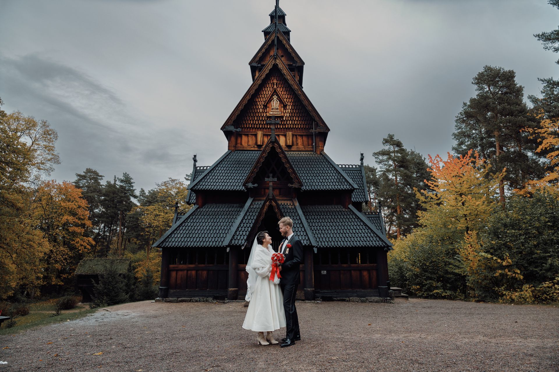 Par i brudeantrekk poserer foran en trekirke med en overskyet himmel i bakgrunnen.