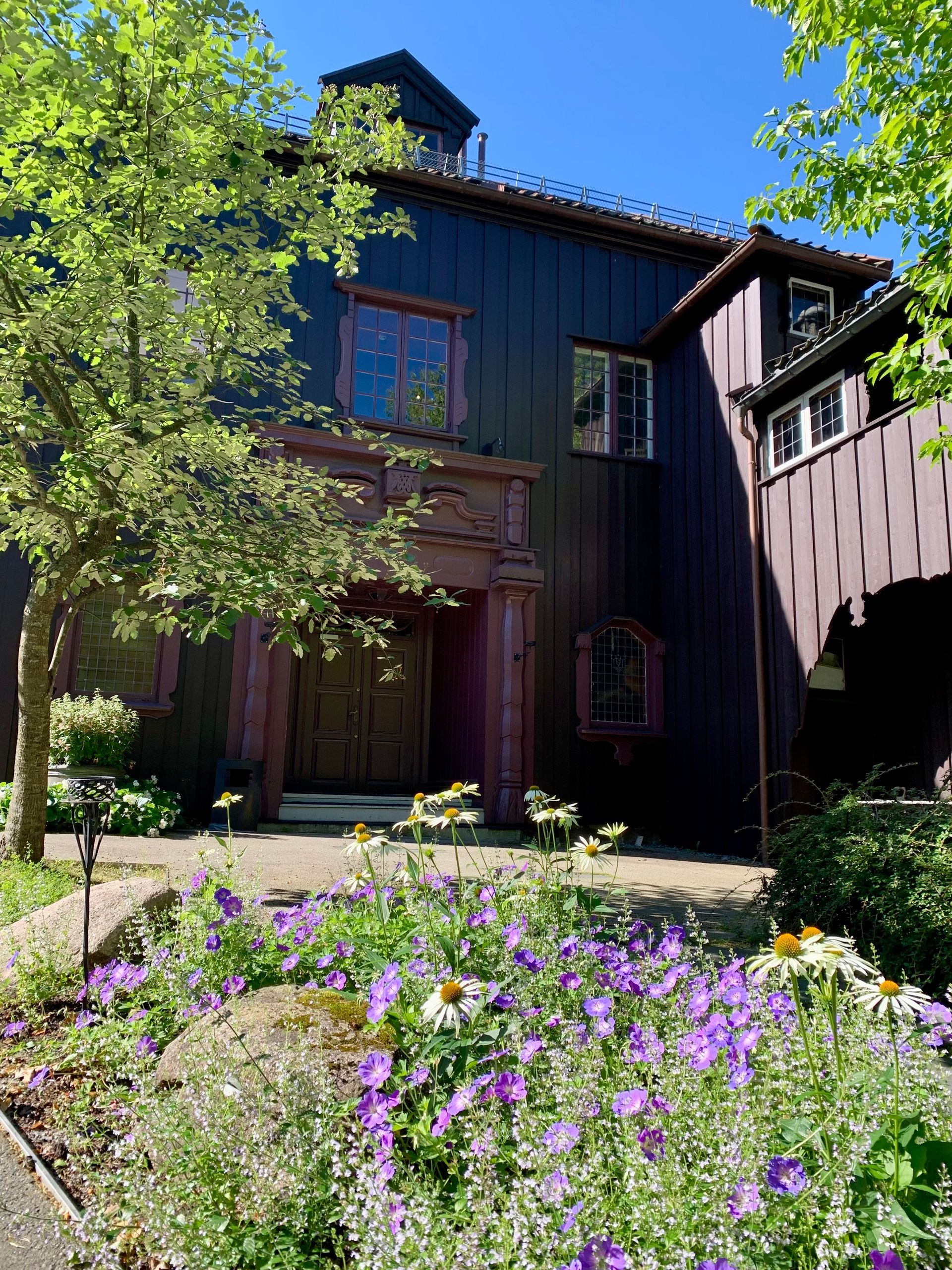 Brun trebygning med rosa listverk, blomster i forgrunnen, blå himmel.