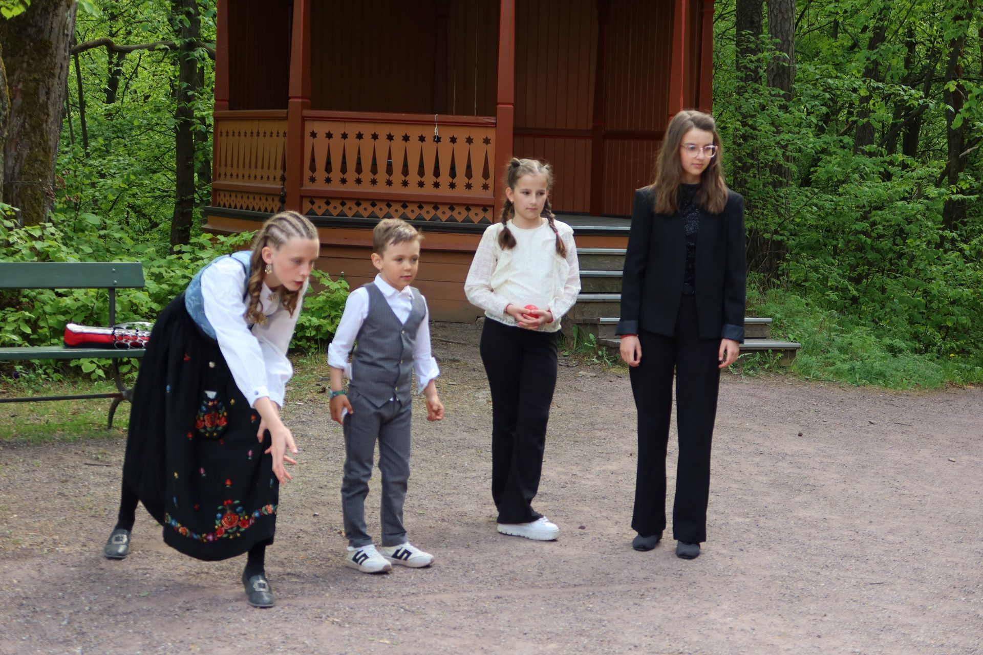 Fire personer utendørs: én i tradisjonell drakt, bøyd; tre andre stående.