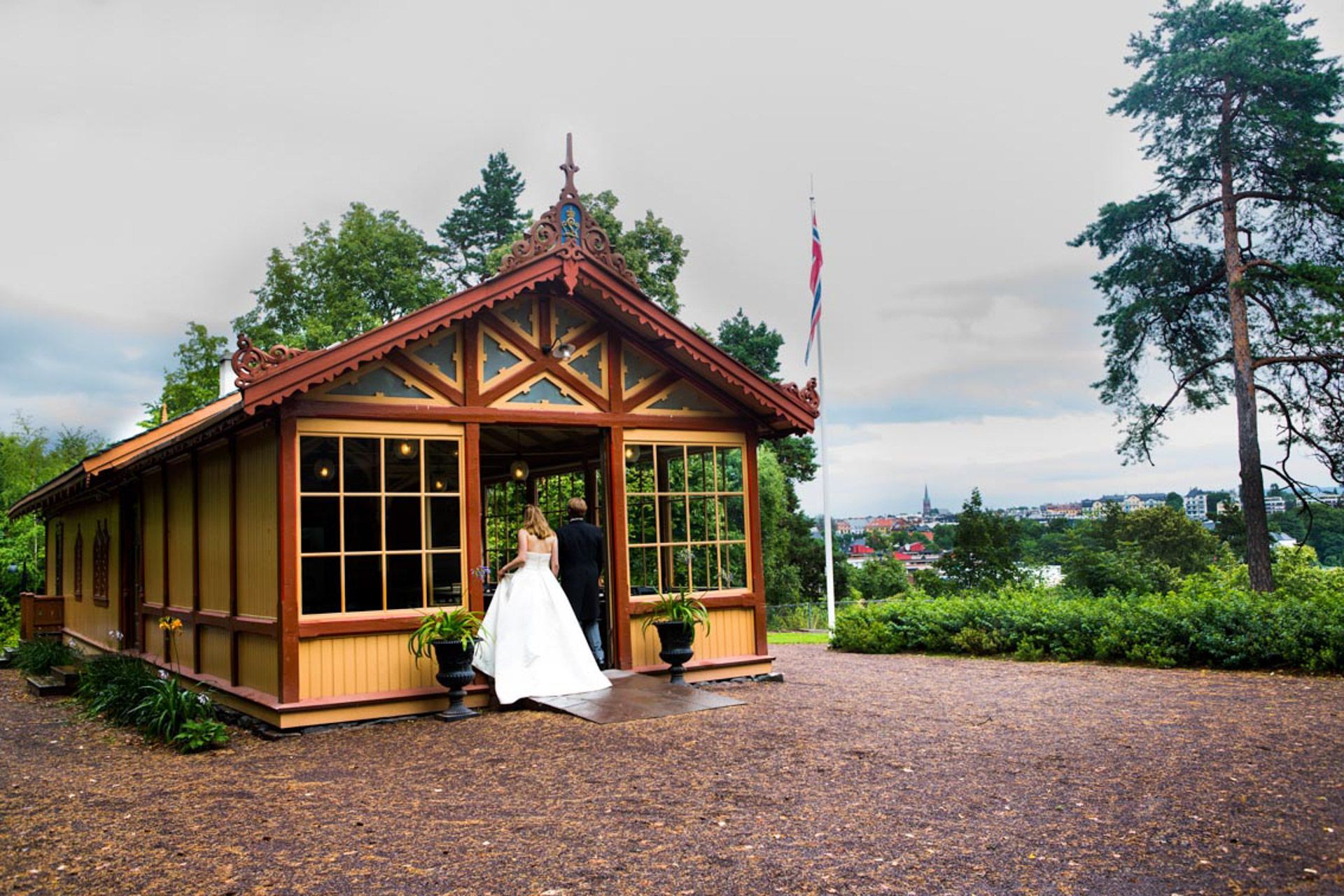 Brud og brudgom går ut av en dekorativ trepaviljong. Brun bygning med store vinduer, beliggende i en park.