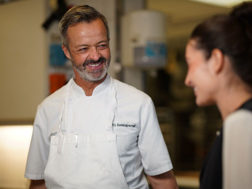 Kokk i hvit uniform smiler, mens han snakker med noen på et kjøkken.