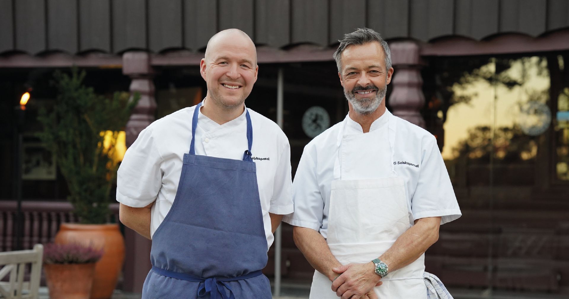 To smilende kokker står foran en bygning. Den ene har på seg hvit frakk og den andre et blått forkle.