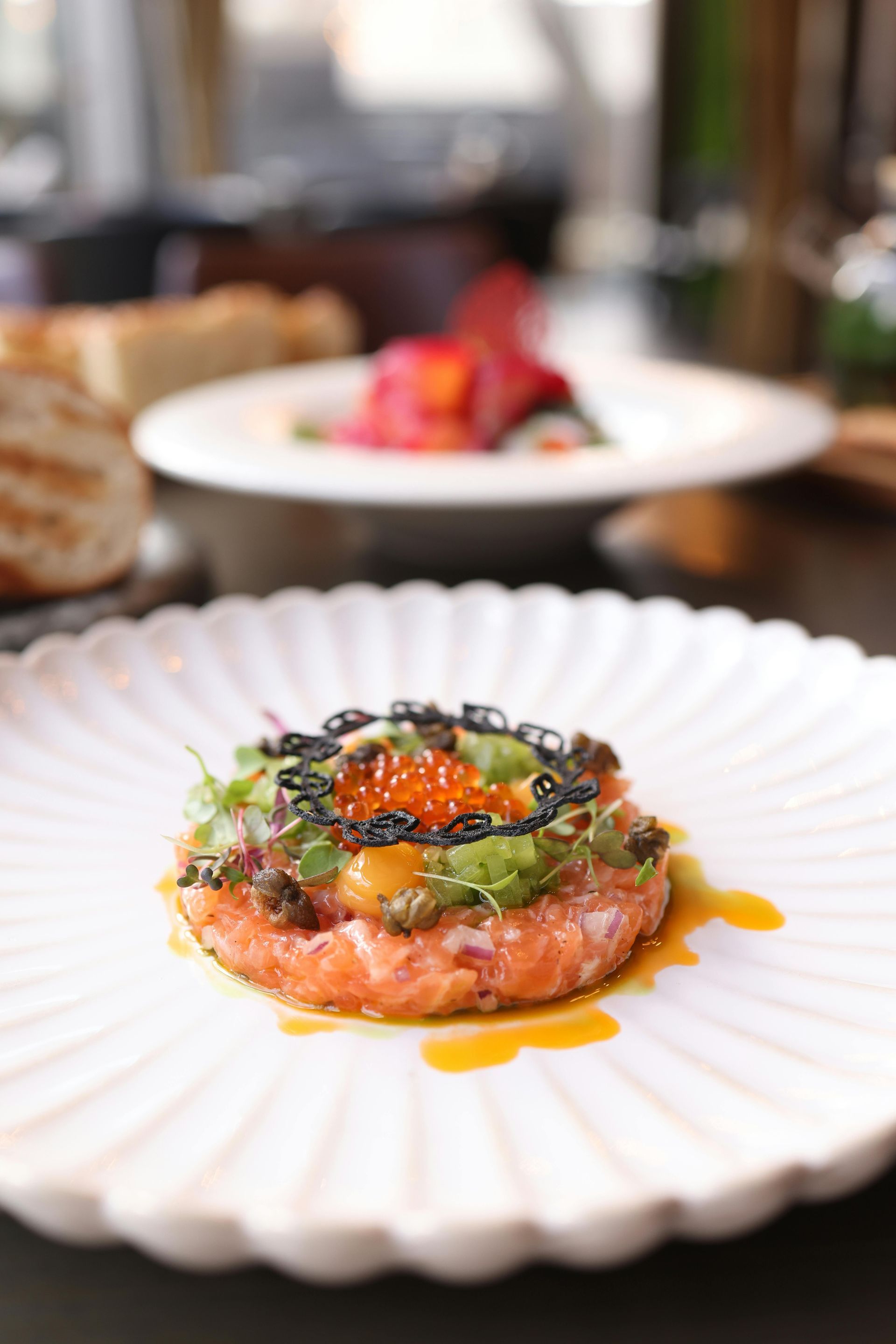 Laksetartar med kaviar og mikrogrønnsaker på en hvit tallerken; andre retter er uskarpe i bakgrunnen.