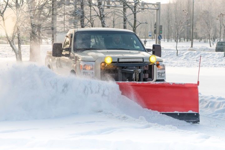 A white pickup truck with a large, bright red snowplow attached to the front clears deep snow from a road.