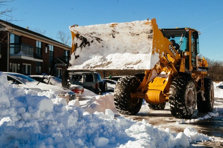 A yellow front-end loader clearing snow from a parking lot near an apartment building on a sunny day.