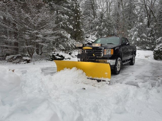 A black pickup truck with a large yellow snowplow attached to the front clears snow from a snowy driveway.