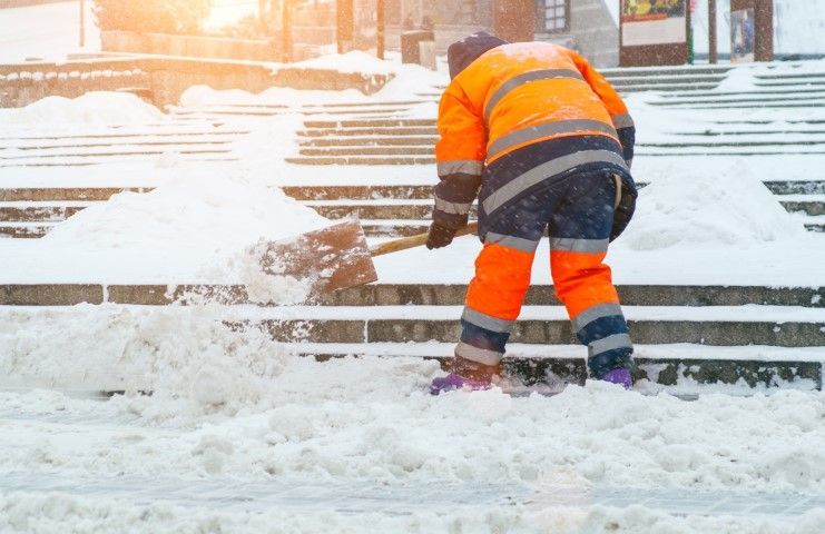 A person in high-visibility orange work clothing uses a shovel to clear snow from stone steps.