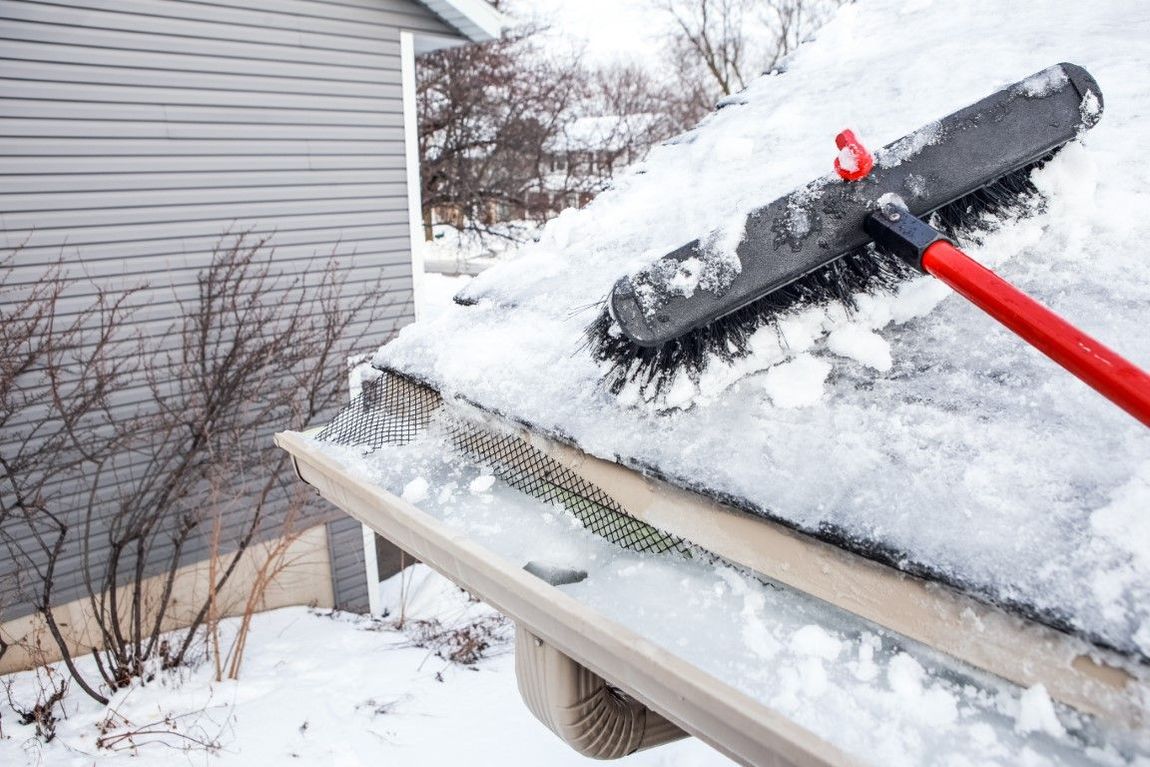 A red-handled roof rake clearing heavy snow from a house roof and gutter.