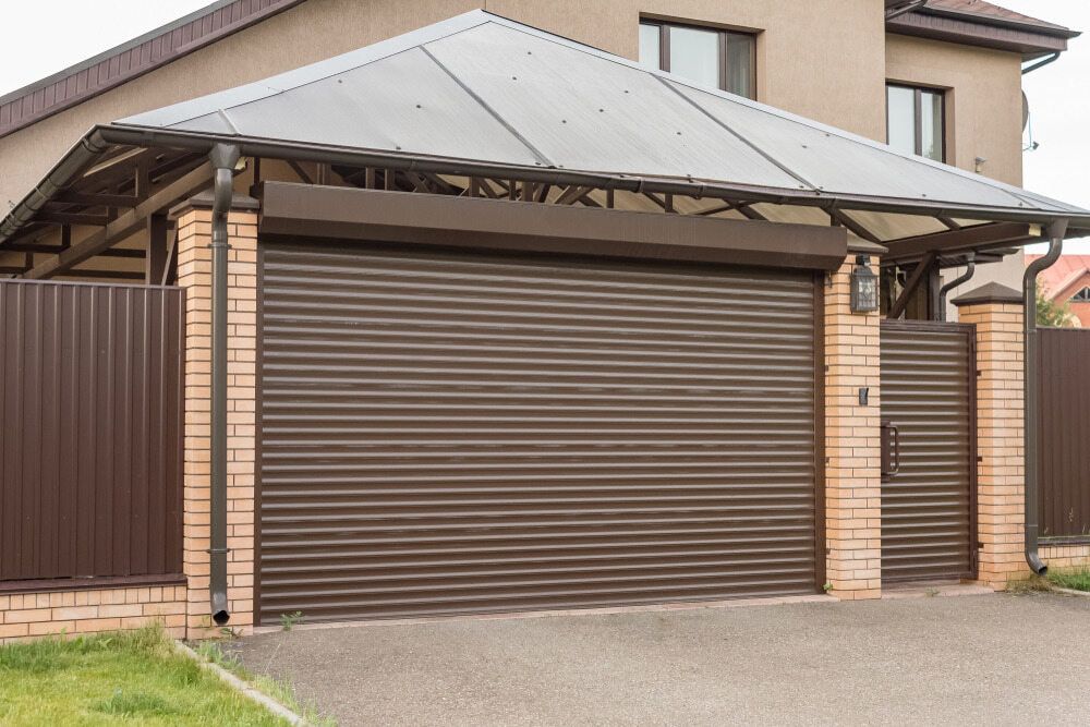 Brown Roll-up Garage Door With Canopy, Between Brick Pillars and Brown Fence — Trade Fencing Supplies in Moolap, VIC