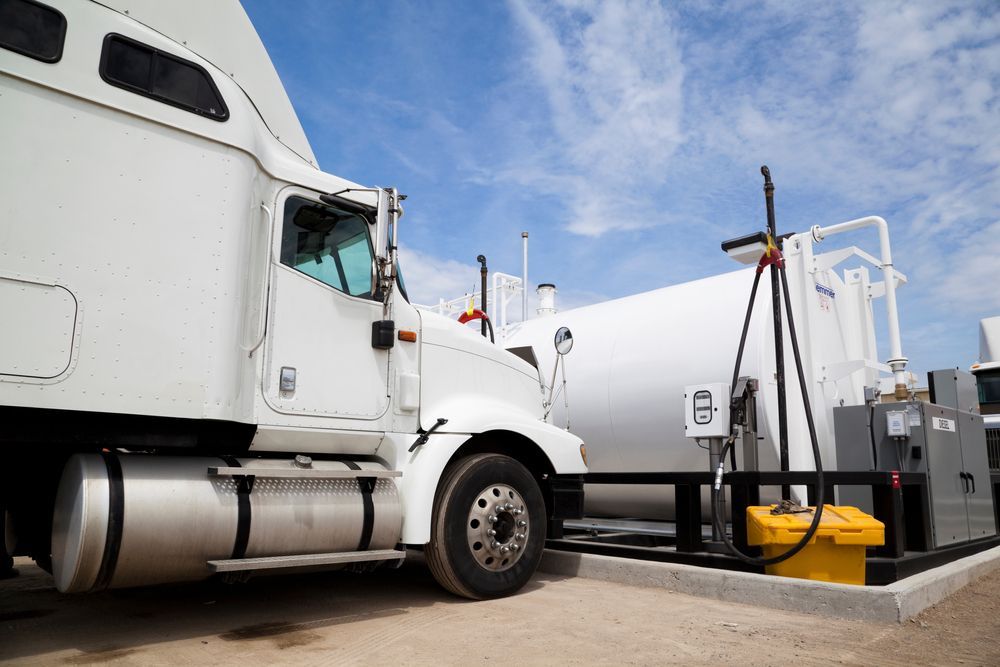 White tanker truck at a fuel terminal beside a large storage tank under a blue sky