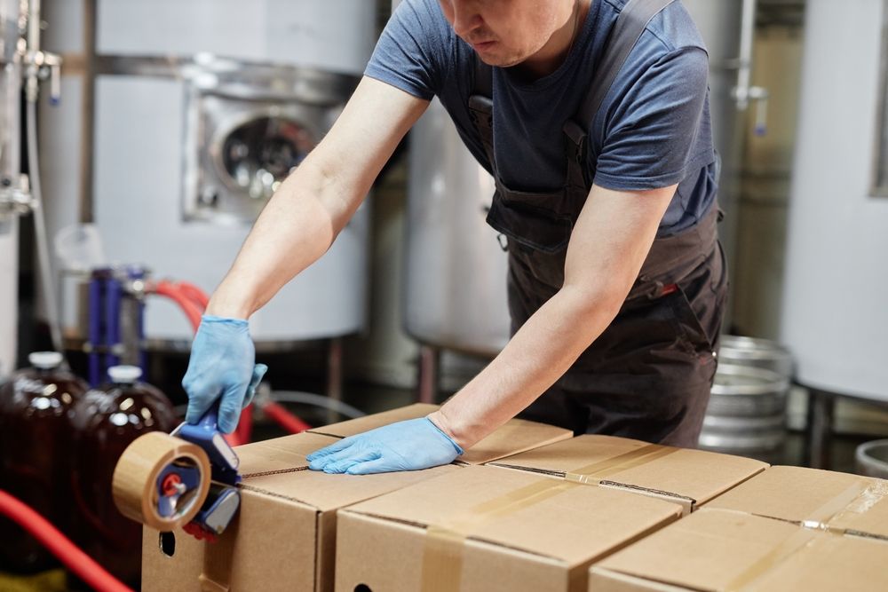 Worker sealing cardboard boxes with tape in food production facility wearing gloves