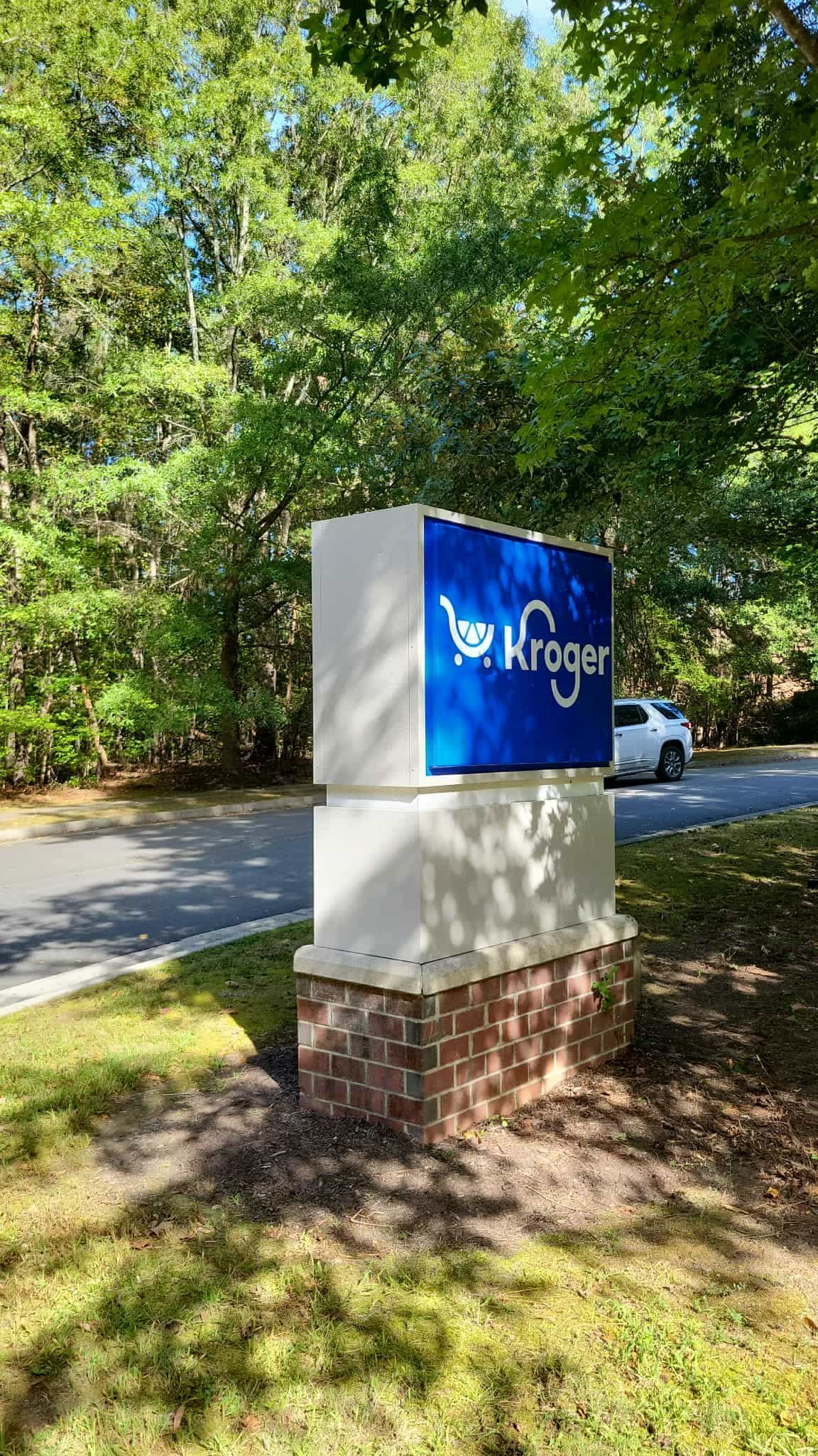 Kroger sign with logo on a white and beige structure, next to a road, with trees in the background.