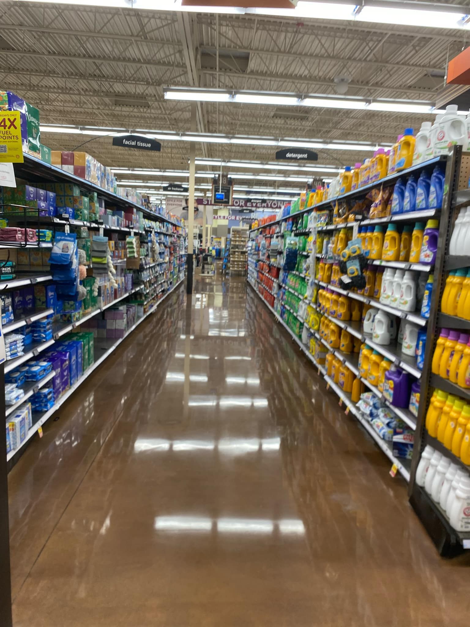 Grocery store aisle with cleaning products on shelves, brown floor, and overhead lights.