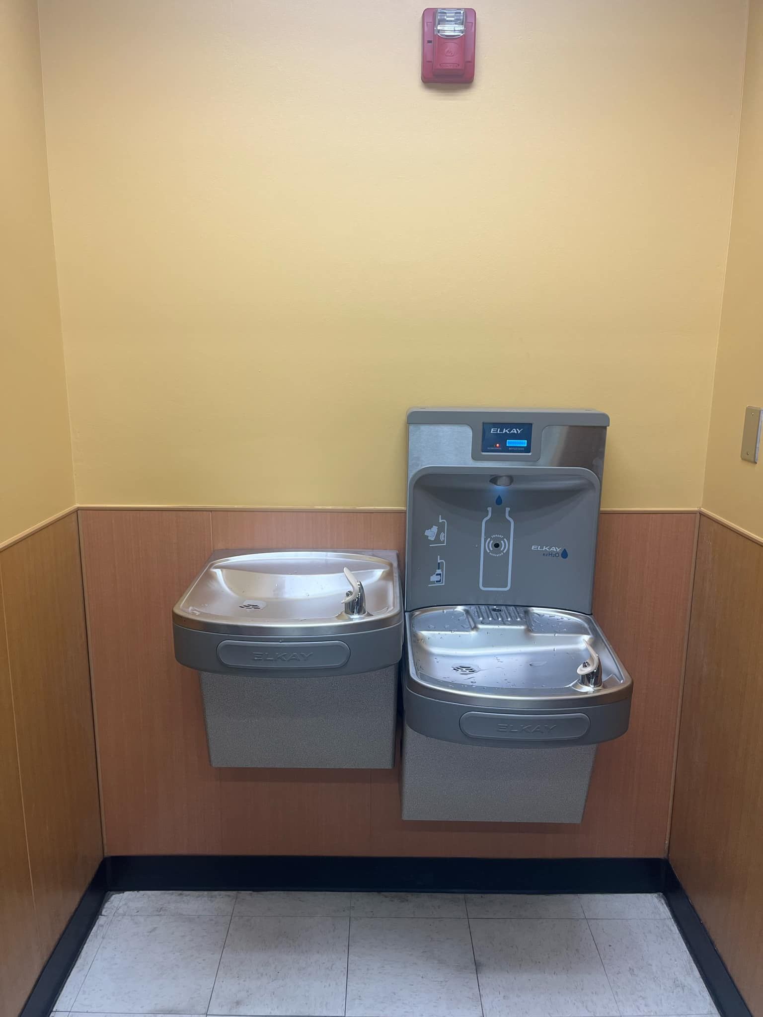 Two water fountains mounted on a wall. One has a bottle filler. Fire alarm above.