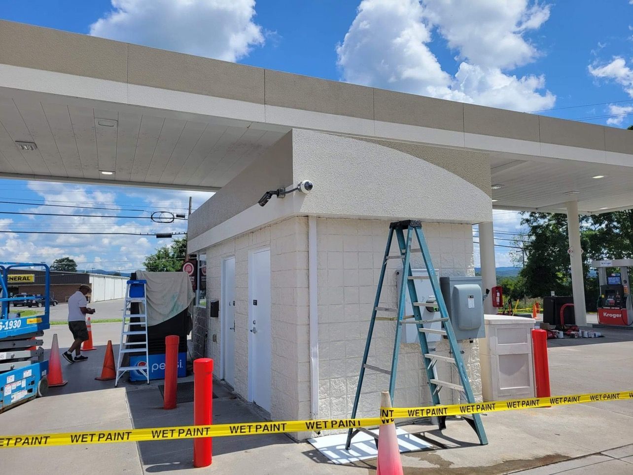 Gas station canopy with a building being painted. Ladder, cones, and caution tape visible. Blue sky.