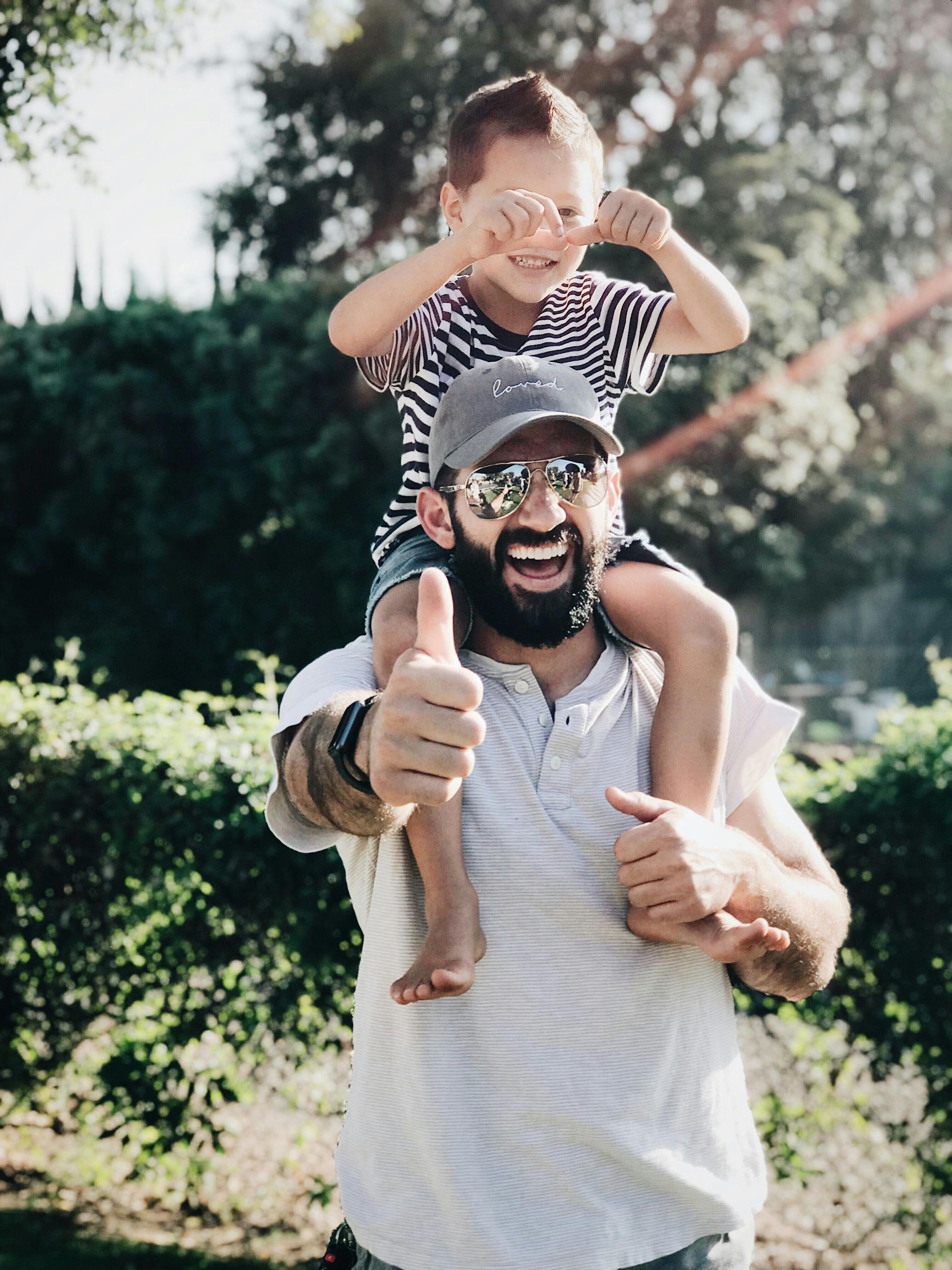 Man giving thumbs up with child on his shoulders; both smiling in a park, sunlight shining.