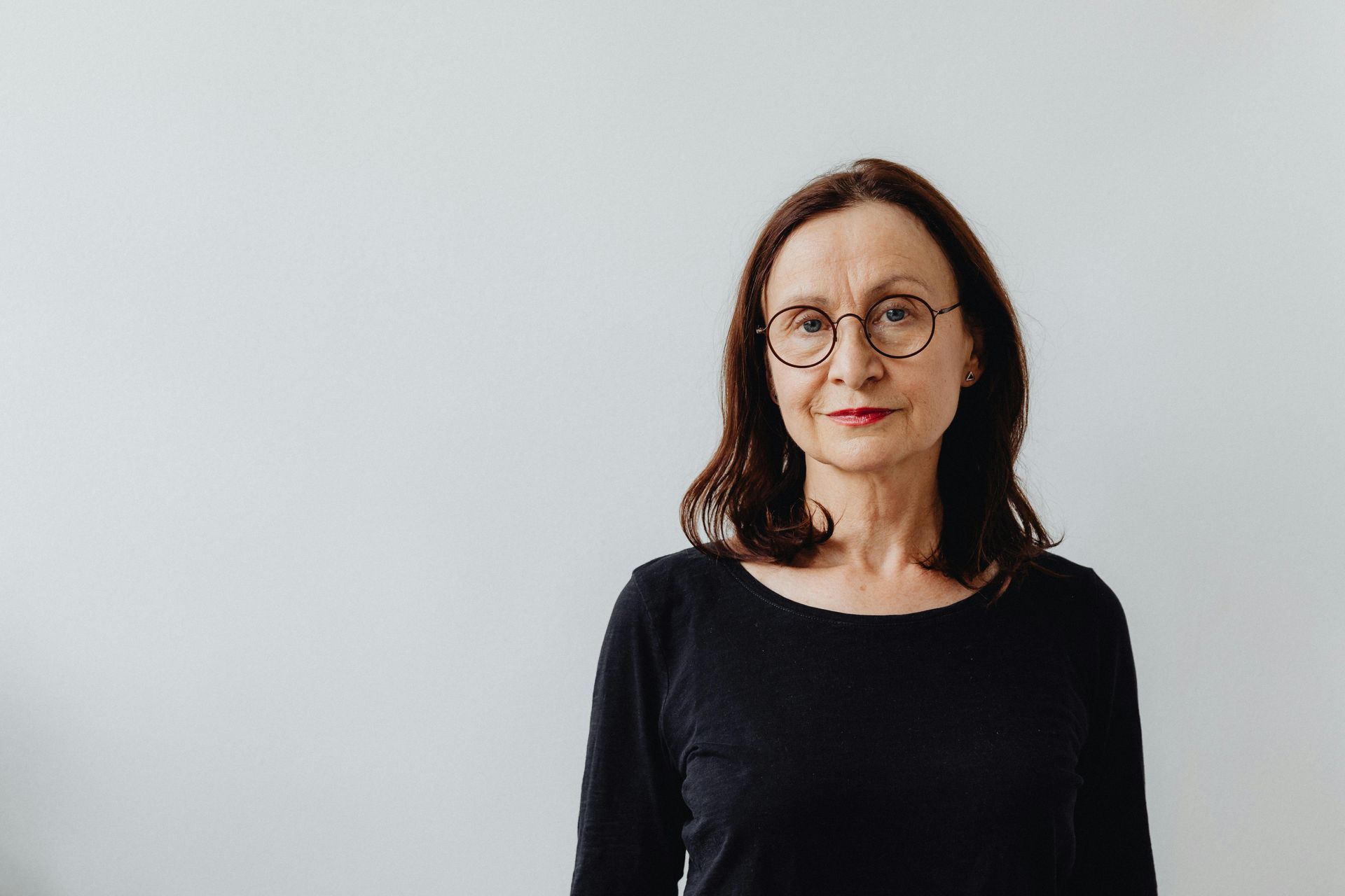 Woman with glasses and dark hair, wearing a black shirt, looking at the camera against a white wall.