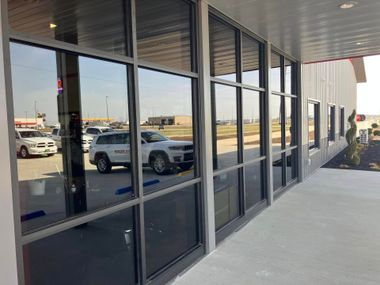 A building exterior with large, dark-tinted glass windows reflecting a parking lot, a white SUV, and a sunny day.