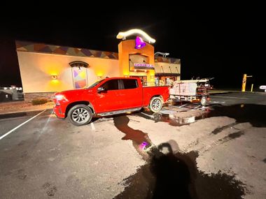 A red pickup truck is parked in front of a Taco Bell at night, towing a utility trailer on a wet, dark parking lot.
