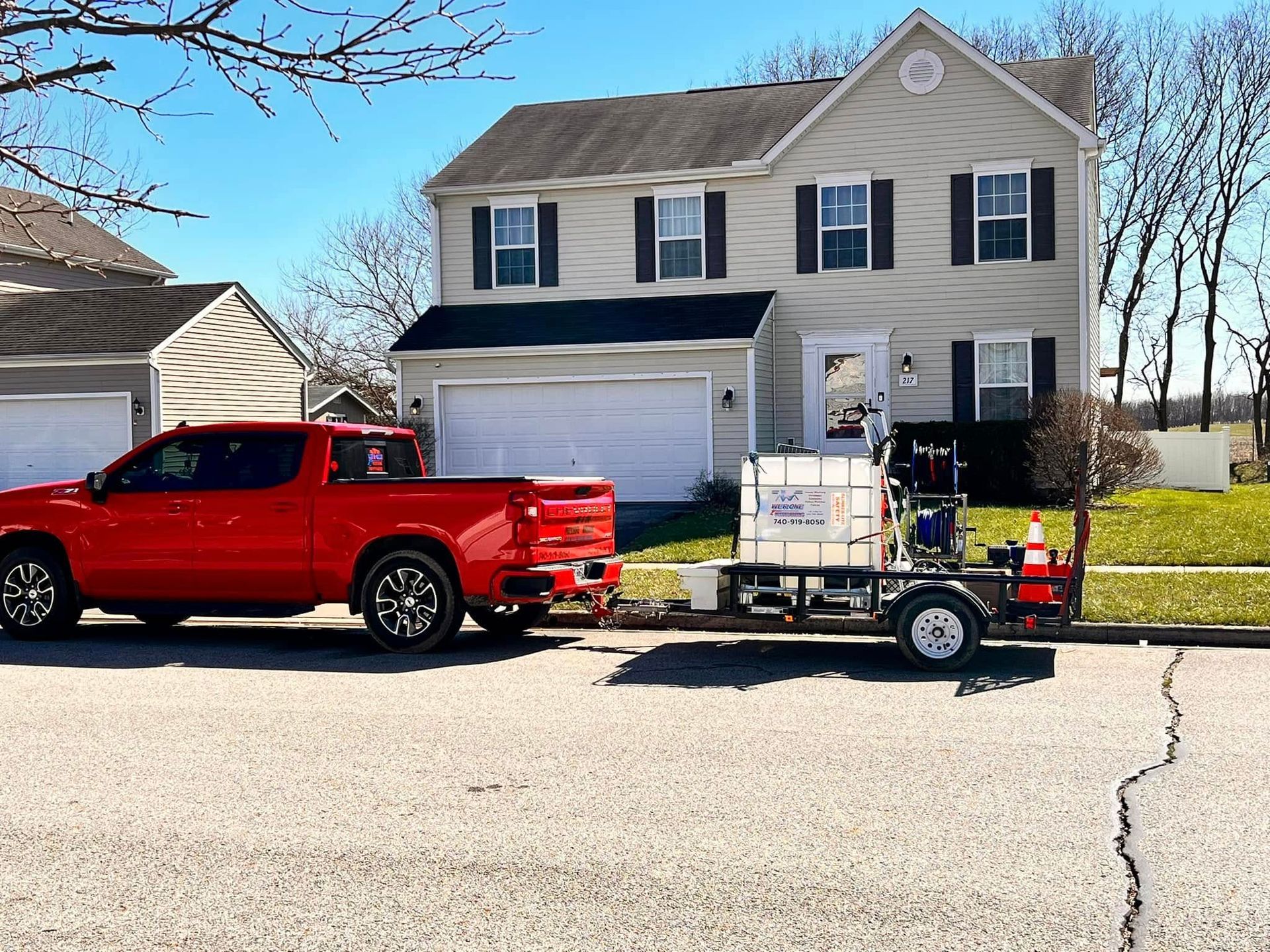 A red pickup truck is parked on a street in front of a suburban house, towing a trailer with a large water tank.