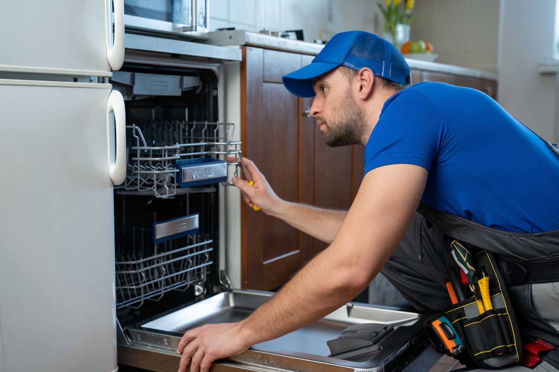 A repairman in a blue shirt and cap works on a dishwasher.