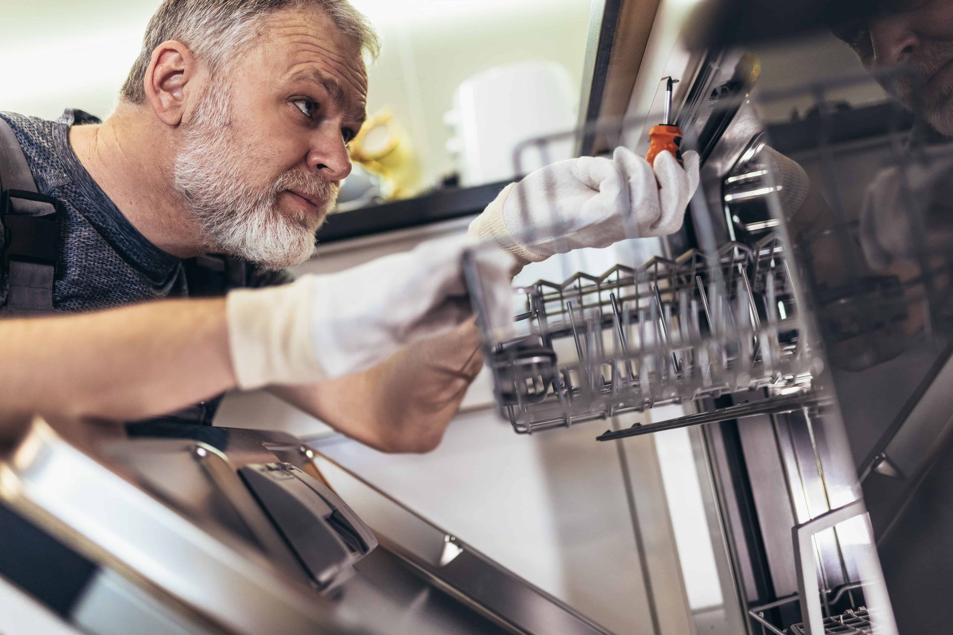 Man in gloves repairing a dishwasher with a screwdriver, looking intently.