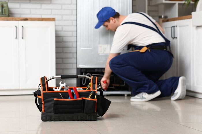 A repairman in a blue uniform working on a refrigerator in a kitchen with a toolbox in front.