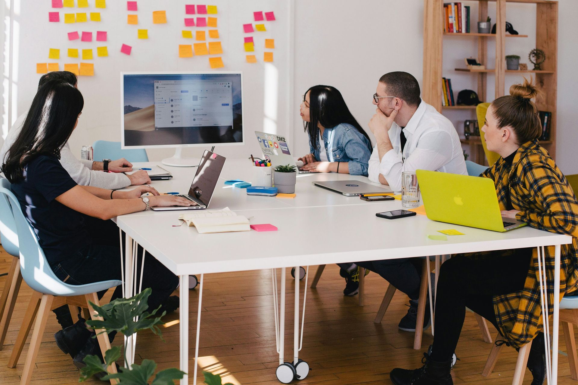 People at a table in a bright office with laptops, a screen, sticky notes, and a plant.