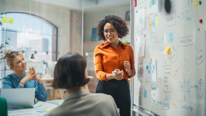 A woman is standing in front of a whiteboard giving a presentation to a group of people.