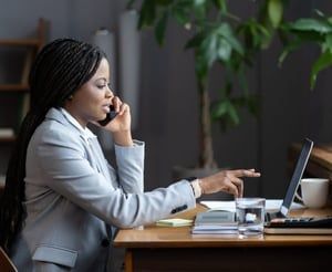 A woman is sitting at a desk talking on a cell phone while using a laptop computer.